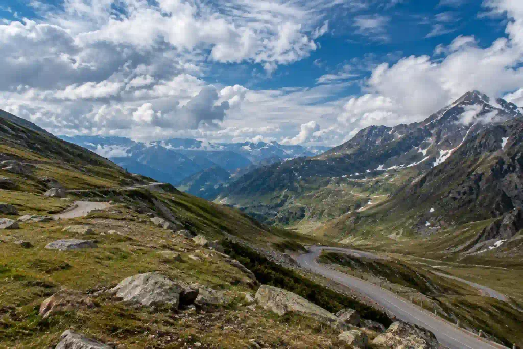 Best Places to Visit in Kashmir 13 High-altitude view of Sinthan Top, Kashmir, with a winding mountain road, rocky alpine terrain, lingering snow and layered Himalayan peaks beneath dramatic clouds.