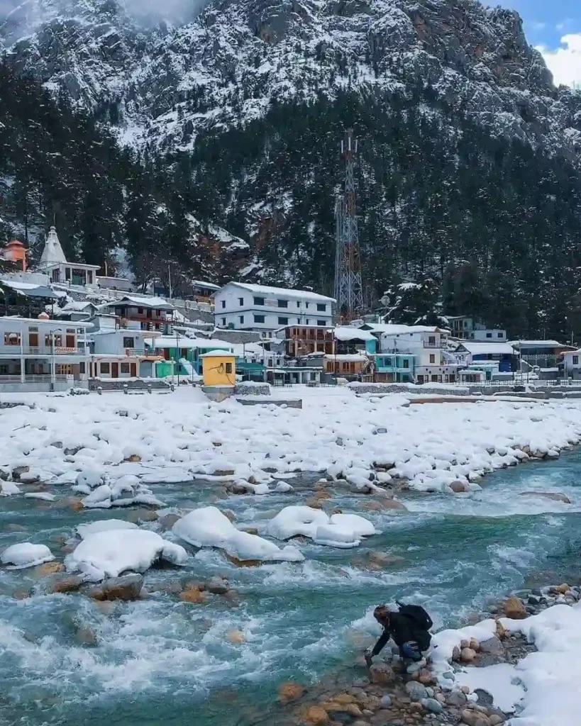Snow-covered Kashmir village beside a flowing river surrounded by snow-capped mountains in winter.
